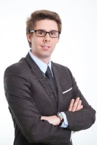 Young man with glasses in a grey suit and tie, arms crossed