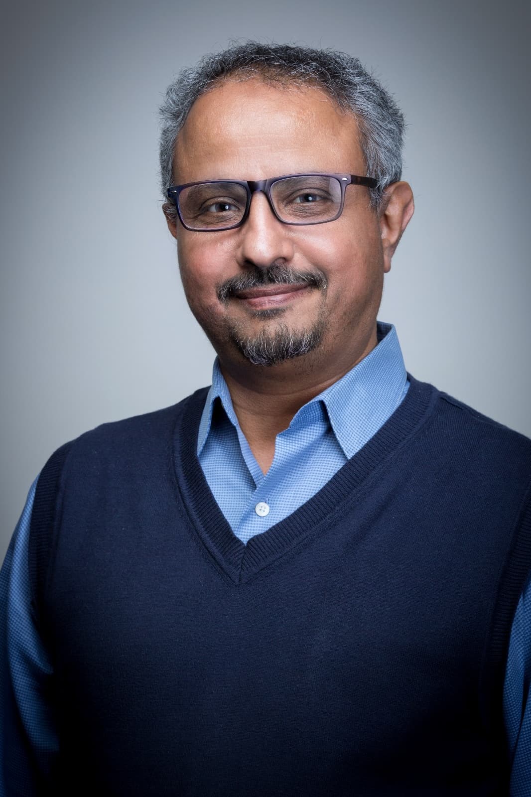 Smiling man with grey hair and glasses in a navy sweater vest over a blue shirt